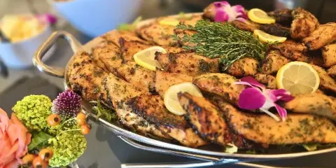 A large silver platter filled with grilled chicken pieces garnished with lemon slices, fresh herbs, and orchids, set on a table with a floral arrangement.