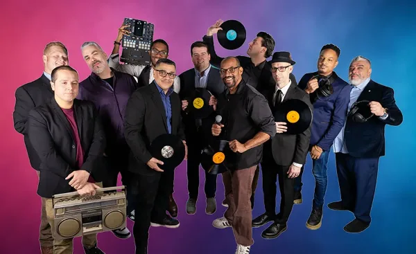 A group of professional DJs and event staff posing with turntables, mixers, and microphones.