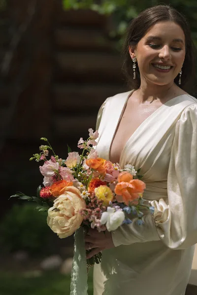 A smiling bride in a cream satin dress holding a colorful bouquet of orange, peach, and white flowers.