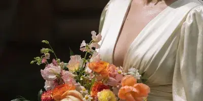 A smiling bride in a cream satin dress holding a colorful bouquet of orange, peach, and white flowers.