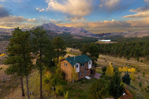 Aerial view of a two-story wooden cabin with a green roof nestled in a mountain landscape with pine trees and a lake in the distance.