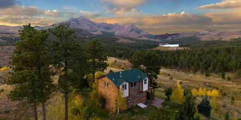 Aerial view of a two-story wooden cabin with a green roof nestled in a mountain landscape with pine trees and a lake in the distance.