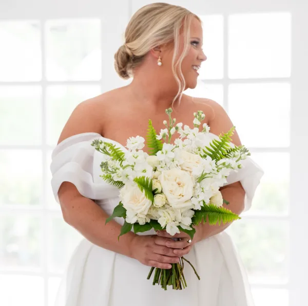 A bride in a white gown holding a bouquet of white roses and greenery.