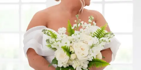 A bride in a white gown holding a bouquet of white roses and greenery.