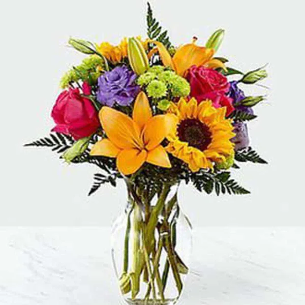 A colorful floral arrangement with sunflowers, orange lilies, and red roses in a clear glass vase.