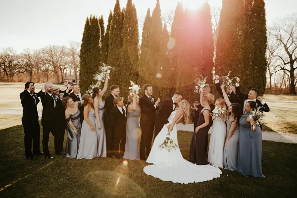 A bride and groom surrounded by their wedding party on a lawn with tall evergreen trees in the background.