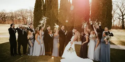 A bride and groom surrounded by their wedding party on a lawn with tall evergreen trees in the background.