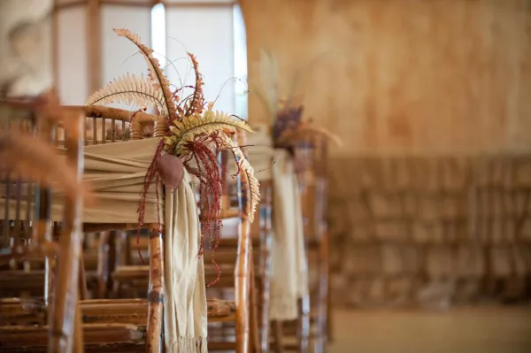 Close-up of a wooden chair decorated with a floral arrangement featuring ferns and dried elements, with a beige fabric sash.