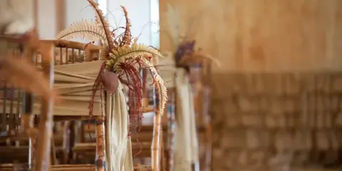 Close-up of a wooden chair decorated with a floral arrangement featuring ferns and dried elements, with a beige fabric sash.