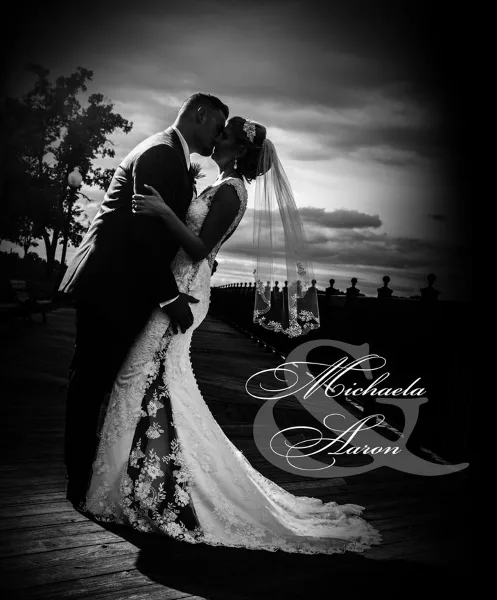 Black and white photograph of a bride and groom kissing on a wooden pier at sunset.