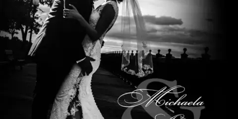 Black and white photograph of a bride and groom kissing on a wooden pier at sunset.