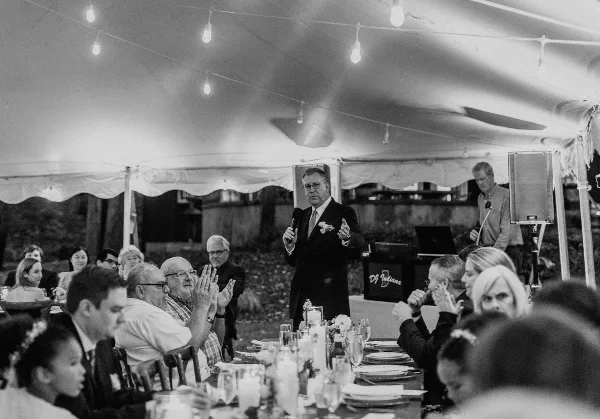 A DJ/MC in a suit addressing a wedding reception audience with a microphone, standing behind a DJ booth with the DJ Indiana logo.
