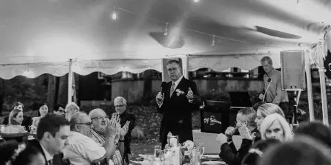 A DJ/MC in a suit addressing a wedding reception audience with a microphone, standing behind a DJ booth with the DJ Indiana logo.