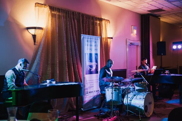 Musicians performing live on stage with pianos and drums in front of a Rock It Man Entertainment banner