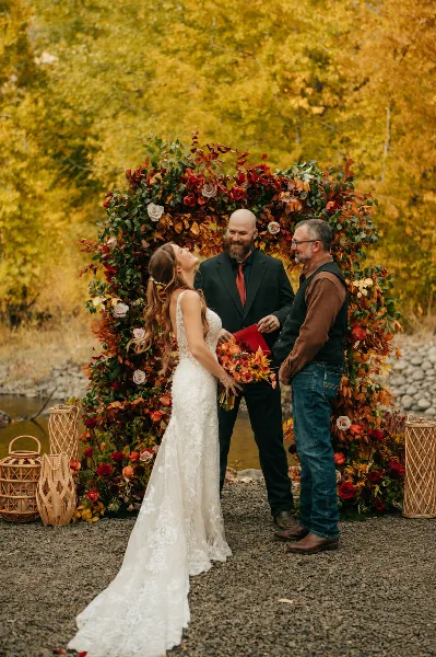 A bride and groom exchanging vows under a floral arch beside a river during an outdoor wedding ceremony.