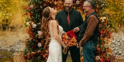 A bride and groom exchanging vows under a floral arch beside a river during an outdoor wedding ceremony.