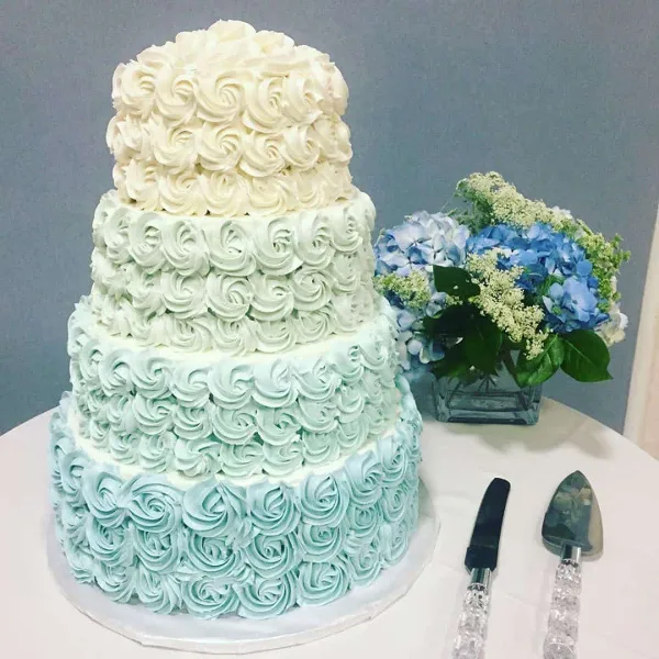 A four-tiered wedding cake with white and light blue rosette frosting, displayed next to a vase of blue hydrangeas and a cake server.