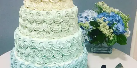 A four-tiered wedding cake with white and light blue rosette frosting, displayed next to a vase of blue hydrangeas and a cake server.