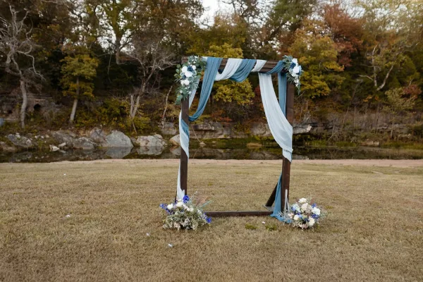 A wooden wedding arch decorated with blue and white flowers and fabric, situated on a grassy lawn near a creek and wooded area.