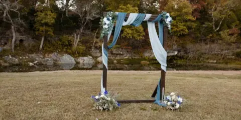 A wooden wedding arch decorated with blue and white flowers and fabric, situated on a grassy lawn near a creek and wooded area.