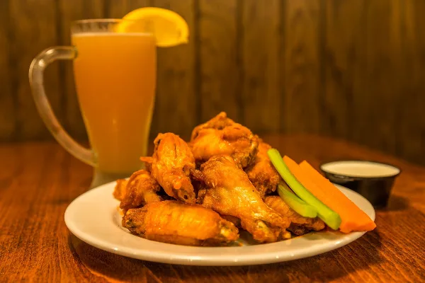 A plate of fried chicken wings with celery and carrot sticks and a small dipping sauce, served with a glass of beer on a wooden table.