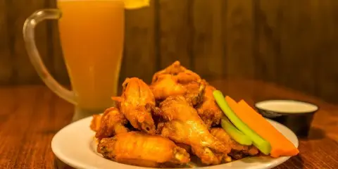 A plate of fried chicken wings with celery and carrot sticks and a small dipping sauce, served with a glass of beer on a wooden table.