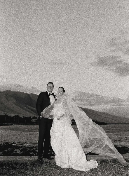 A black and white photograph of a bride and groom standing on a rocky beach with a mountain range in the background.