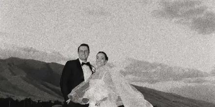 A black and white photograph of a bride and groom standing on a rocky beach with a mountain range in the background.