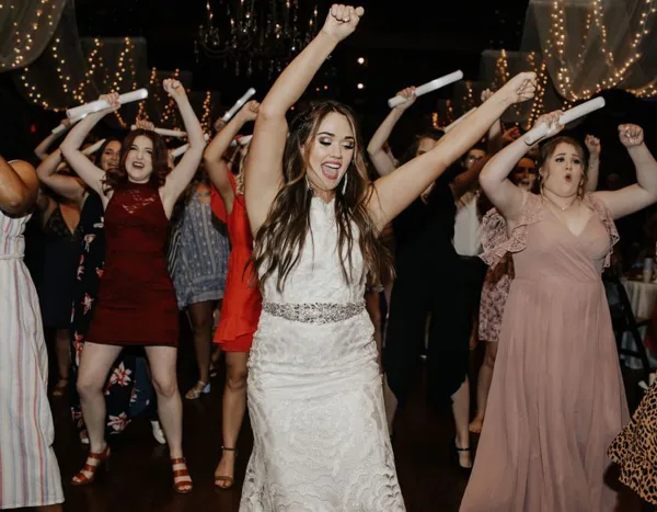 A bride in a white lace dress dancing with her wedding party at a reception, holding glow sticks.