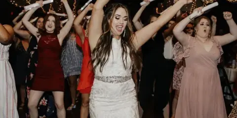 A bride in a white lace dress dancing with her wedding party at a reception, holding glow sticks.