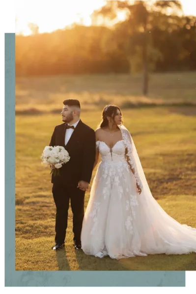A bride and groom standing in a grassy field at sunset, with the bride holding a bouquet of white flowers.