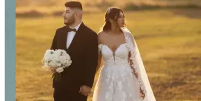 A bride and groom standing in a grassy field at sunset, with the bride holding a bouquet of white flowers.