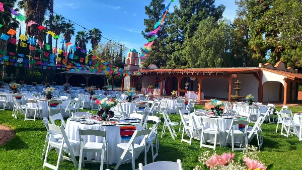Outdoor wedding venue with set tables, white chairs, and a Spanish-style building under a blue sky.