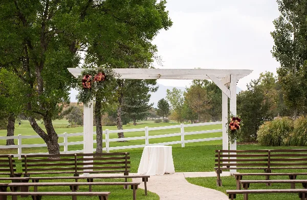 Outdoor wedding ceremony setup with a white pergola, floral arrangements, and wooden benches on a green lawn.