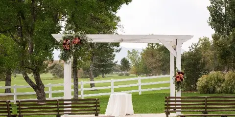 Outdoor wedding ceremony setup with a white pergola, floral arrangements, and wooden benches on a green lawn.