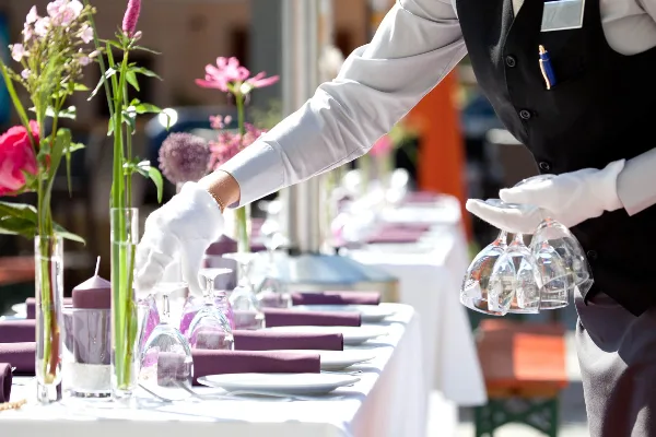 A catering staff member in a black vest and white gloves setting a formal banquet table with flowers and glassware.