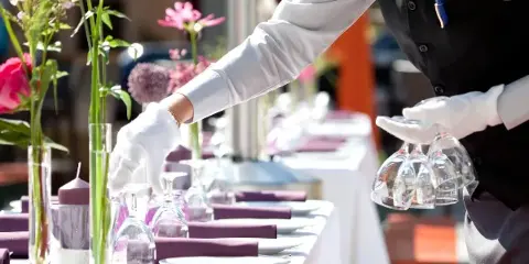 A catering staff member in a black vest and white gloves setting a formal banquet table with flowers and glassware.