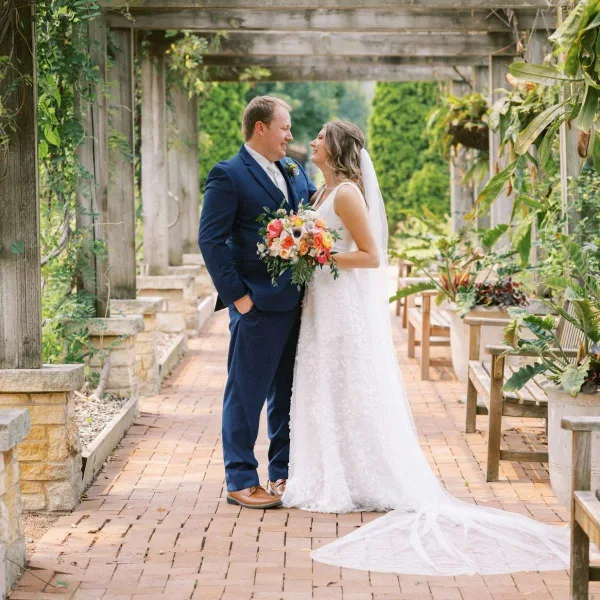 Bride and groom standing together under a wooden arbor in a garden.