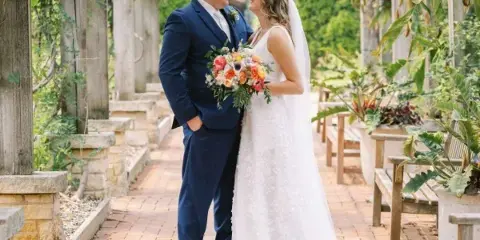Bride and groom standing together under a wooden arbor in a garden.