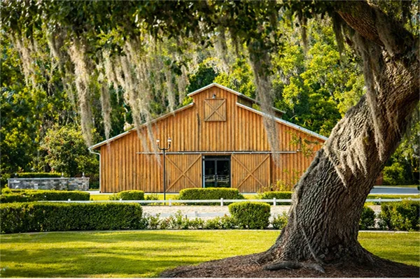 Exterior view of a wooden event barn at an estate, surrounded by manicured hedges and large live oak trees with Spanish moss.
