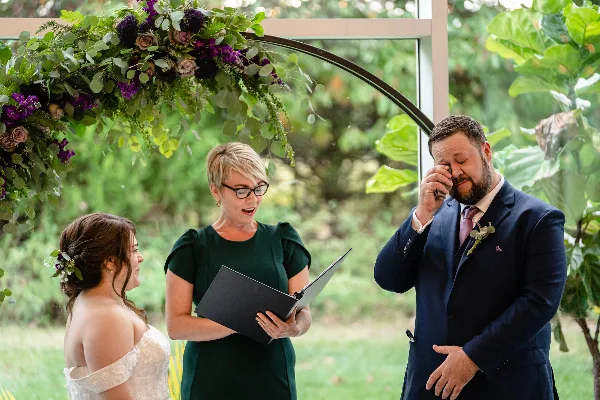 A female officiant in a green dress reading from a folder to a bride and groom during an outdoor wedding ceremony.