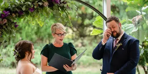 A female officiant in a green dress reading from a folder to a bride and groom during an outdoor wedding ceremony.