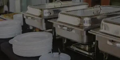 A catering buffet table with stainless steel chafing dishes, stacks of white plates, and trays of prepared food.