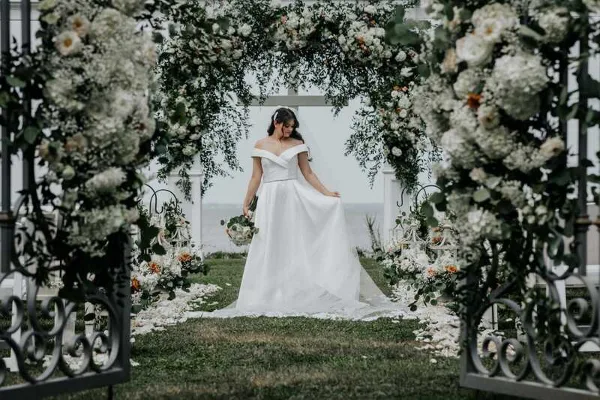 A bride in a white wedding dress standing under a floral archway with a view of the water.