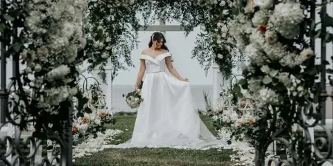 A bride in a white wedding dress standing under a floral archway with a view of the water.