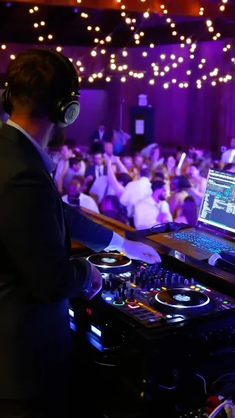 A professional DJ wearing headphones operates a mixing console and laptop at a wedding reception with a crowded dance floor and string lights in the background.