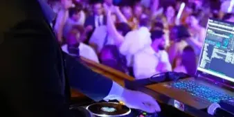 A professional DJ wearing headphones operates a mixing console and laptop at a wedding reception with a crowded dance floor and string lights in the background.