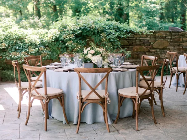 Outdoor event table set with white tablecloth, wooden cross-back chairs, glassware, and floral centerpiece on a stone patio.