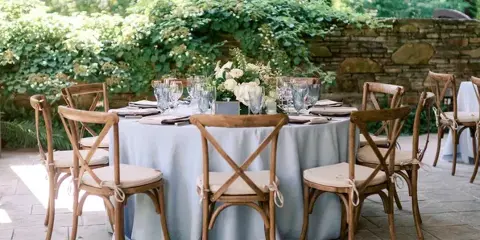 Outdoor event table set with white tablecloth, wooden cross-back chairs, glassware, and floral centerpiece on a stone patio.