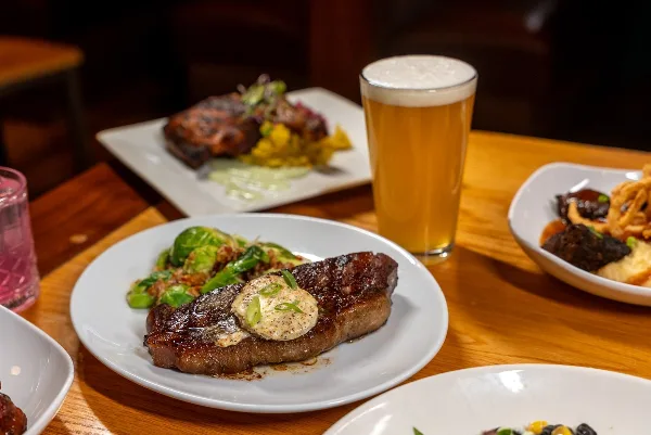 A table setting featuring a plate of steak with a butter topping, a glass of beer, and other prepared dishes.